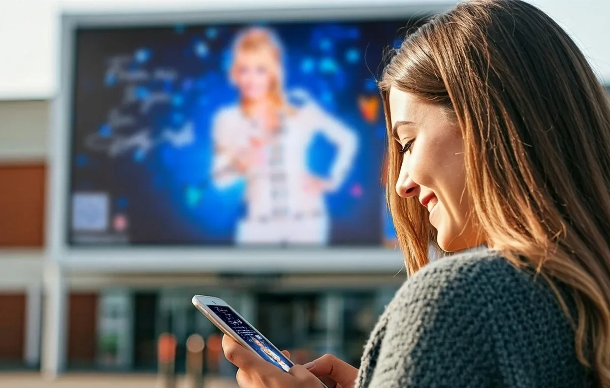 A smiling woman looking at a phone with a graphic matching on a large TV screen in the background