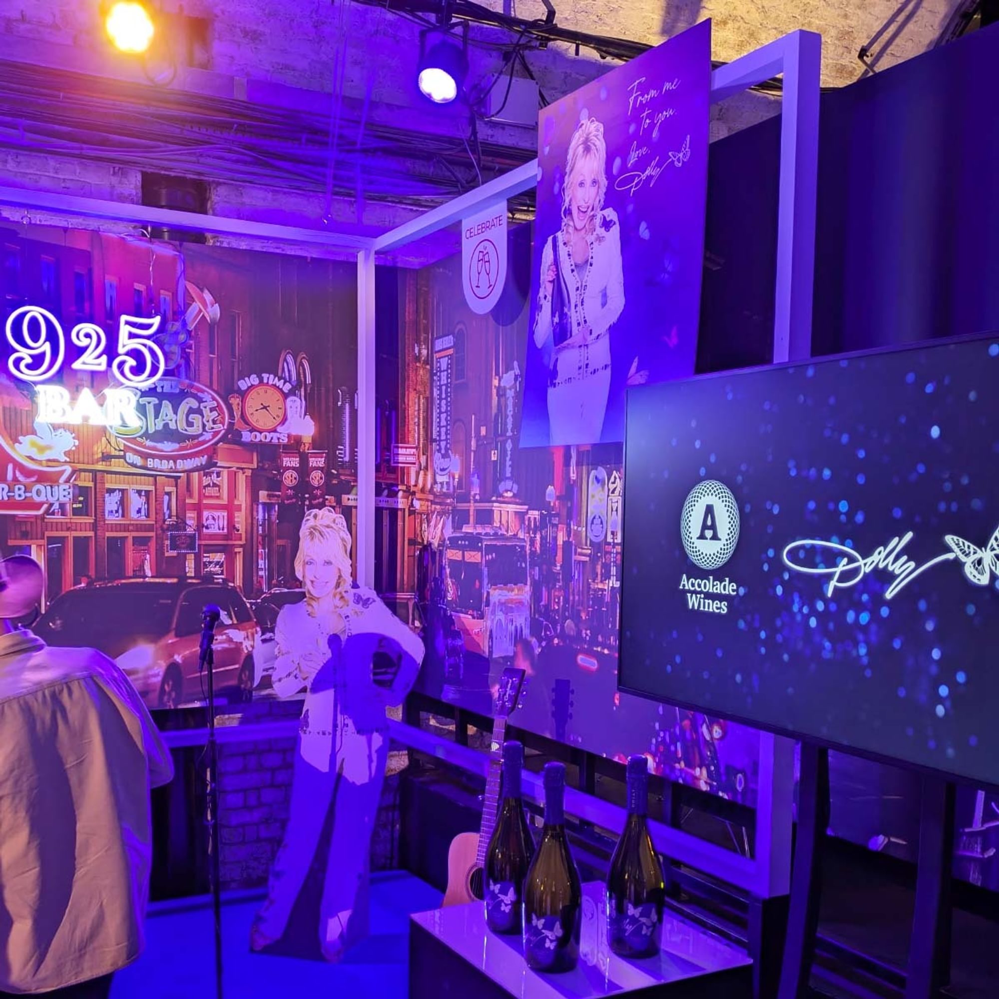 Dolly Parton 9 2 5 bar scene. A card standee on a stage with guitar, mic and neon light. Prints and Wines in the background.