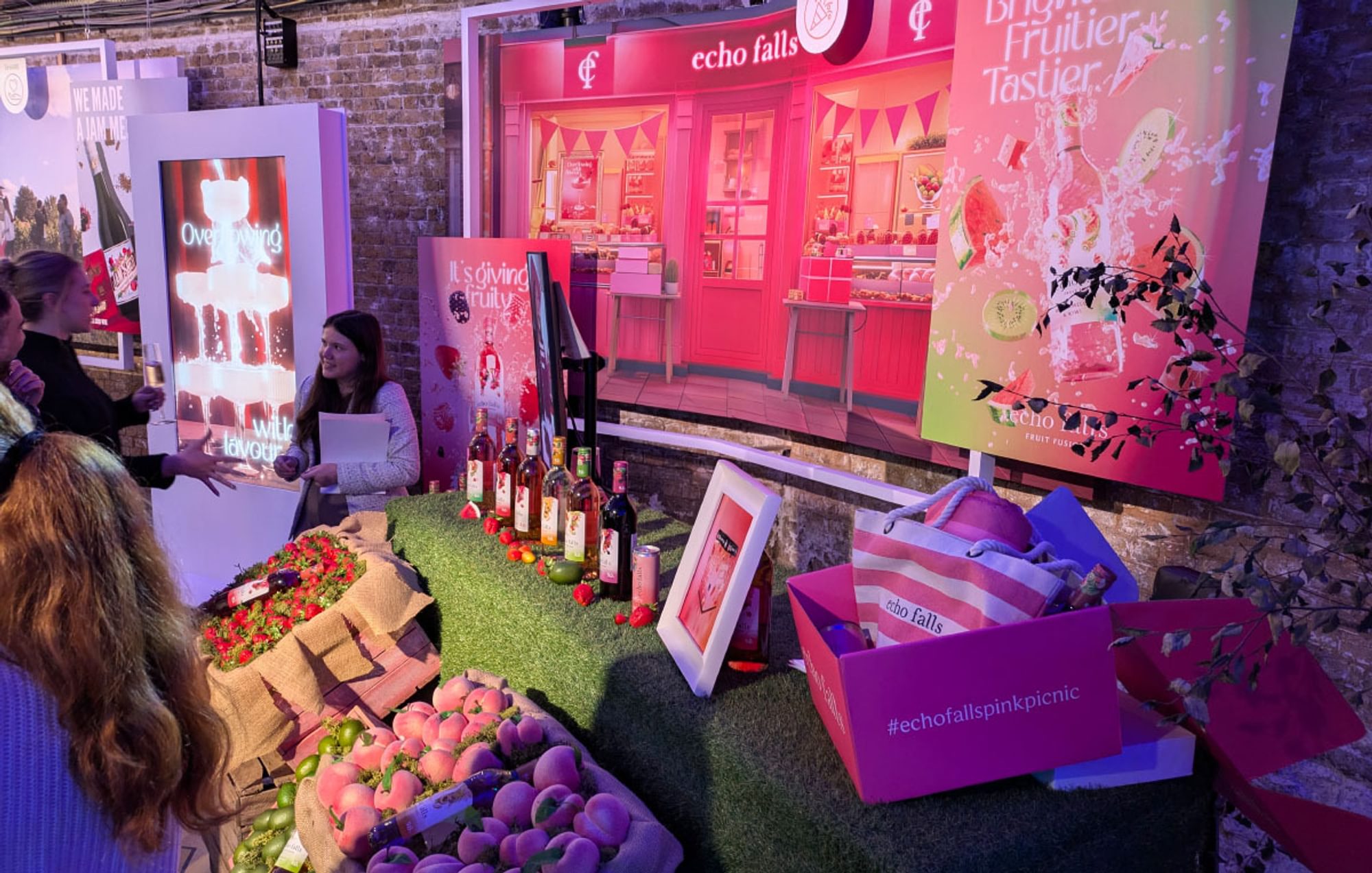 A busy, staffed Echo Falls wine display stand with boxes of fruits, bottles of wine and posters, digital displays and more.
