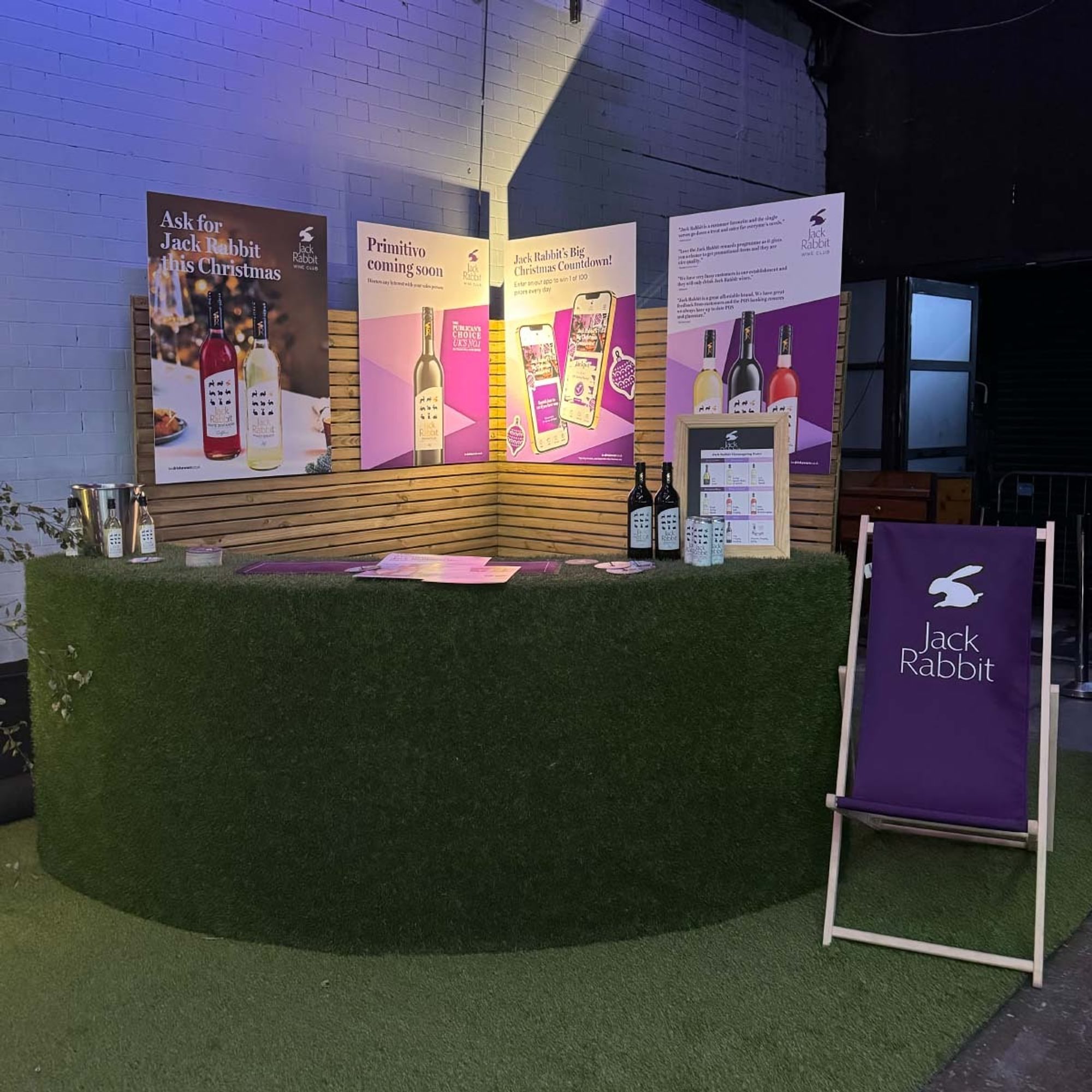 Outdoor bar styled area with large branded posters. Shown are bottles, coasters and barware, with ready to drink wine cans.