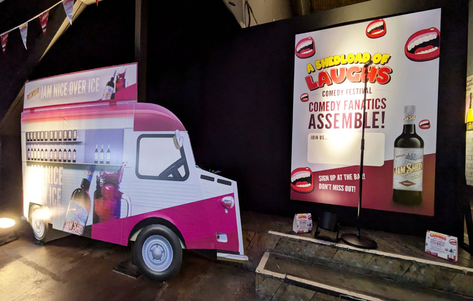 Stand up comedy zone featuring a large van-shaped cardboard standee, mic stand, and large background poster promoting the event.