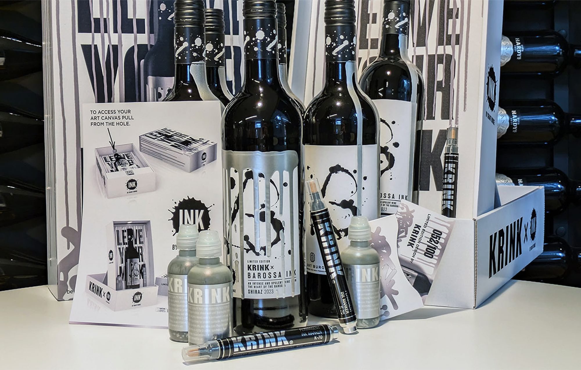 A selection of labelled wine and ink bottles, pens, leaflets and branded boxes against a wall of wine bottles in racks.