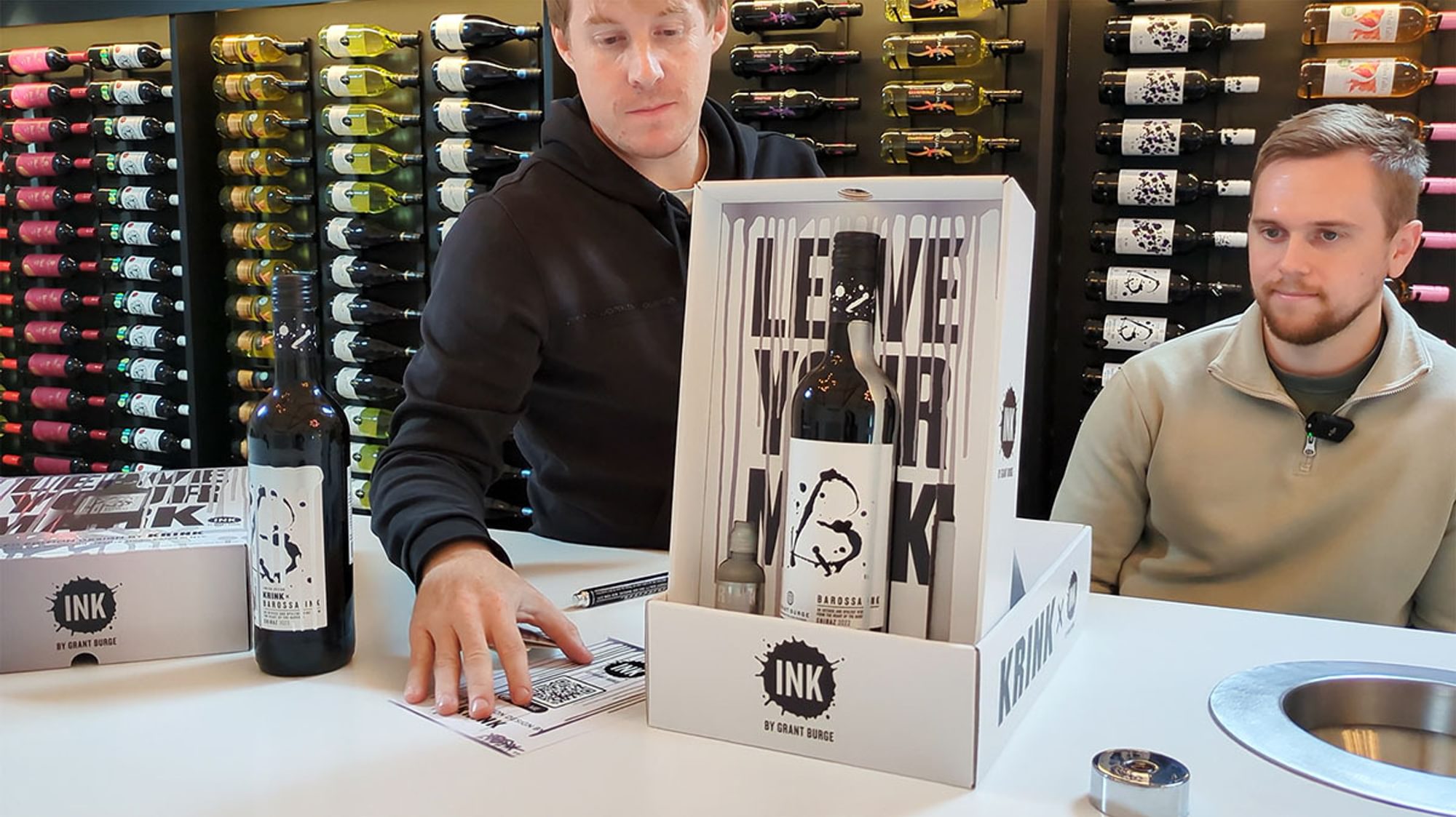 Two men sit in front of a rack filled with hundreds of wine bottles as they show off the box and its contents.